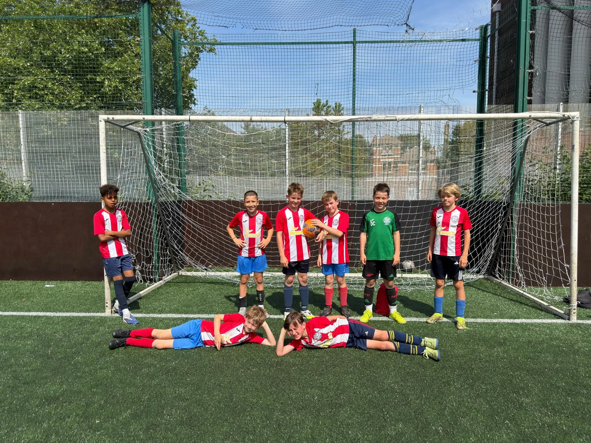 Group of young boys in soccer uniforms standing and lying on artificial turf soccer field, with goal net and tall green fences behind them.
