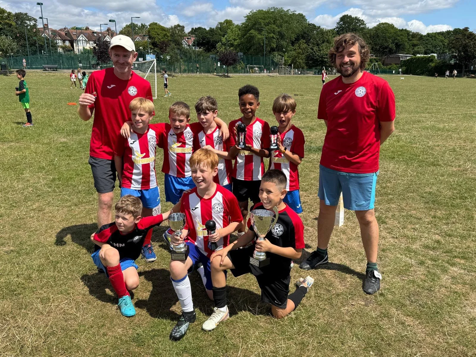 A youth soccer team with two coaches celebrating on a grassy field after a game or tournament. The children are holding trophies and medals, wearing red and white striped jerseys, and smiling. The coaches stand on either side of the team, one in a re