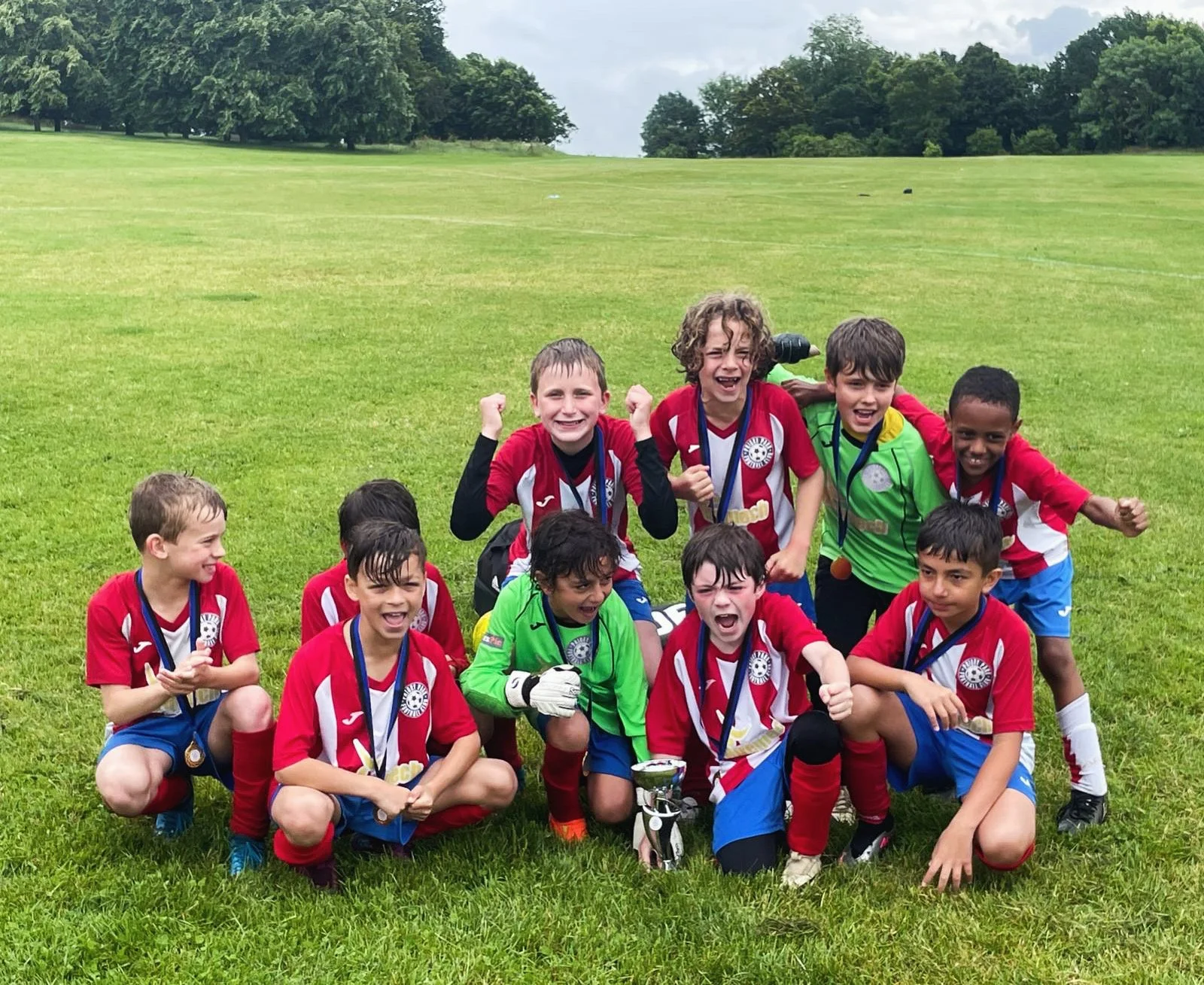 A group of young boys and one girl in soccer uniforms celebrating on a grassy field after winning a game, with medals around their necks and some holding a trophy.