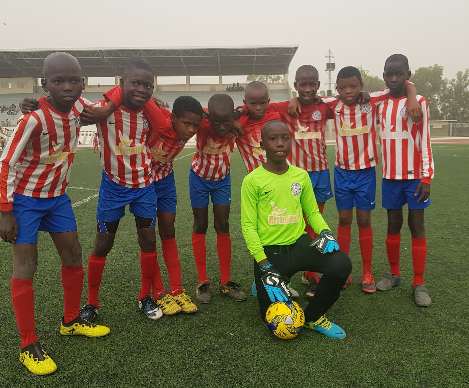 A group of young soccer players, mostly boys, standing together on a soccer field. They are wearing red and white striped jerseys, blue shorts, and red socks. One boy is dressed as a goalkeeper in a bright green shirt and black shorts, kneeling in fr
