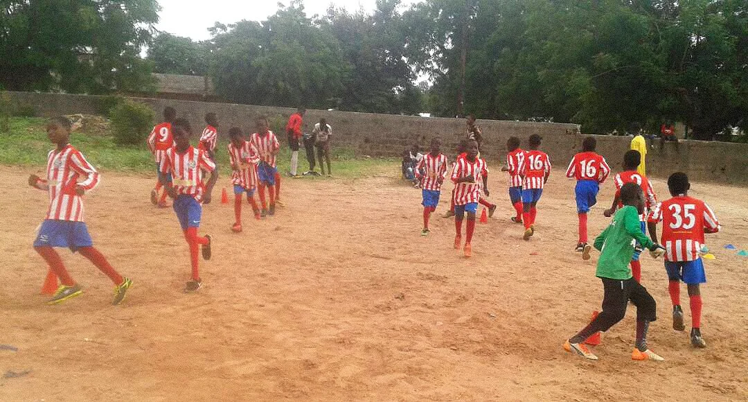 Young soccer players in red and white striped jerseys and blue shorts warming up or training on a dirt field, with some resting against a stone wall and a few adults watching in the background.