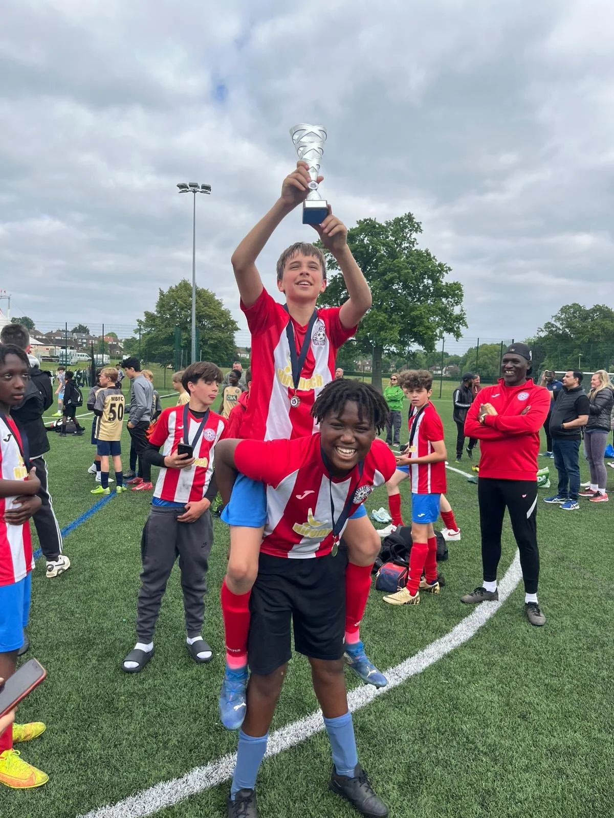 Young soccer players celebrating on the field after a match, with the boy in red and white jersey holding a trophy high in the air.
