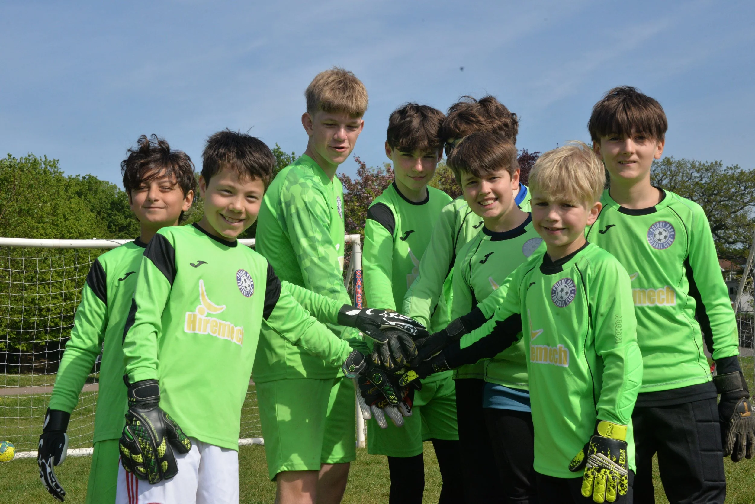 Group of young boys on a soccer field, wearing bright green goalkeeper jerseys, standing in front of a soccer goalpost, with trees and a blue sky in the background.
