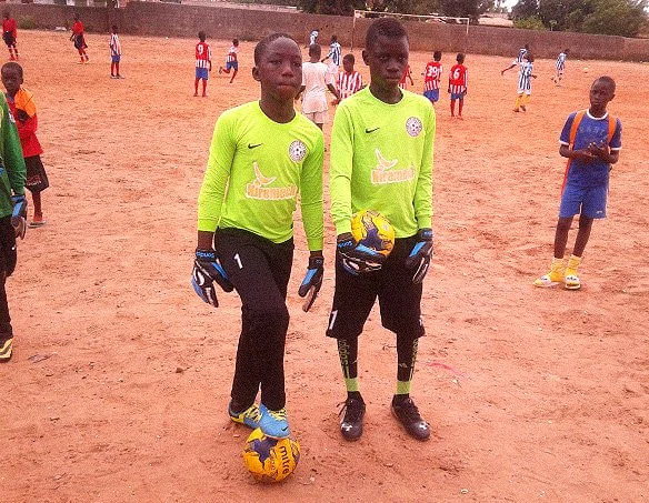 Two young boys in bright green goalkeeper jerseys and black pants standing on a dirt soccer field, one holding a yellow soccer ball and the other resting a foot on a soccer ball. Several children and adults are in the background, some wearing sports uniforms.