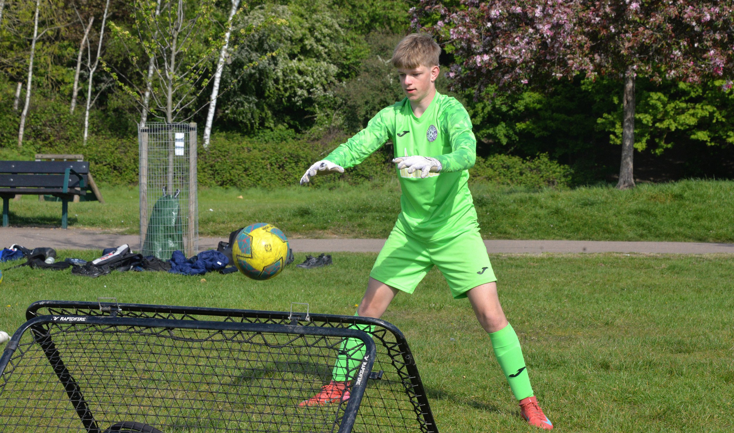 A young male soccer goalkeeper wearing bright green uniform practicing in a park with trees and blooming flowers in the background. He is preparing to kick a yellow and blue soccer ball, standing behind a small black goal.