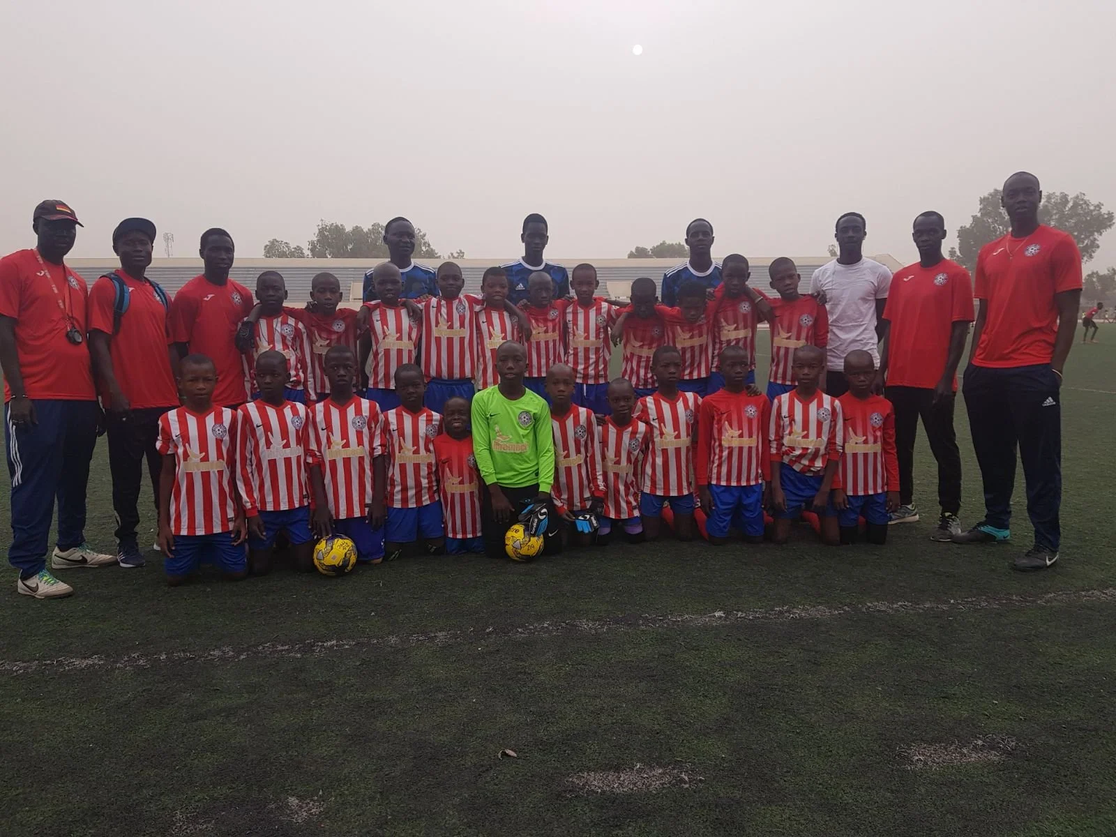 A group of young boys and their coaches on a soccer field, dressed in red and white striped jerseys and blue shorts, with some holding soccer balls and a few wearing goalkeeper jerseys, posing for a team photo.