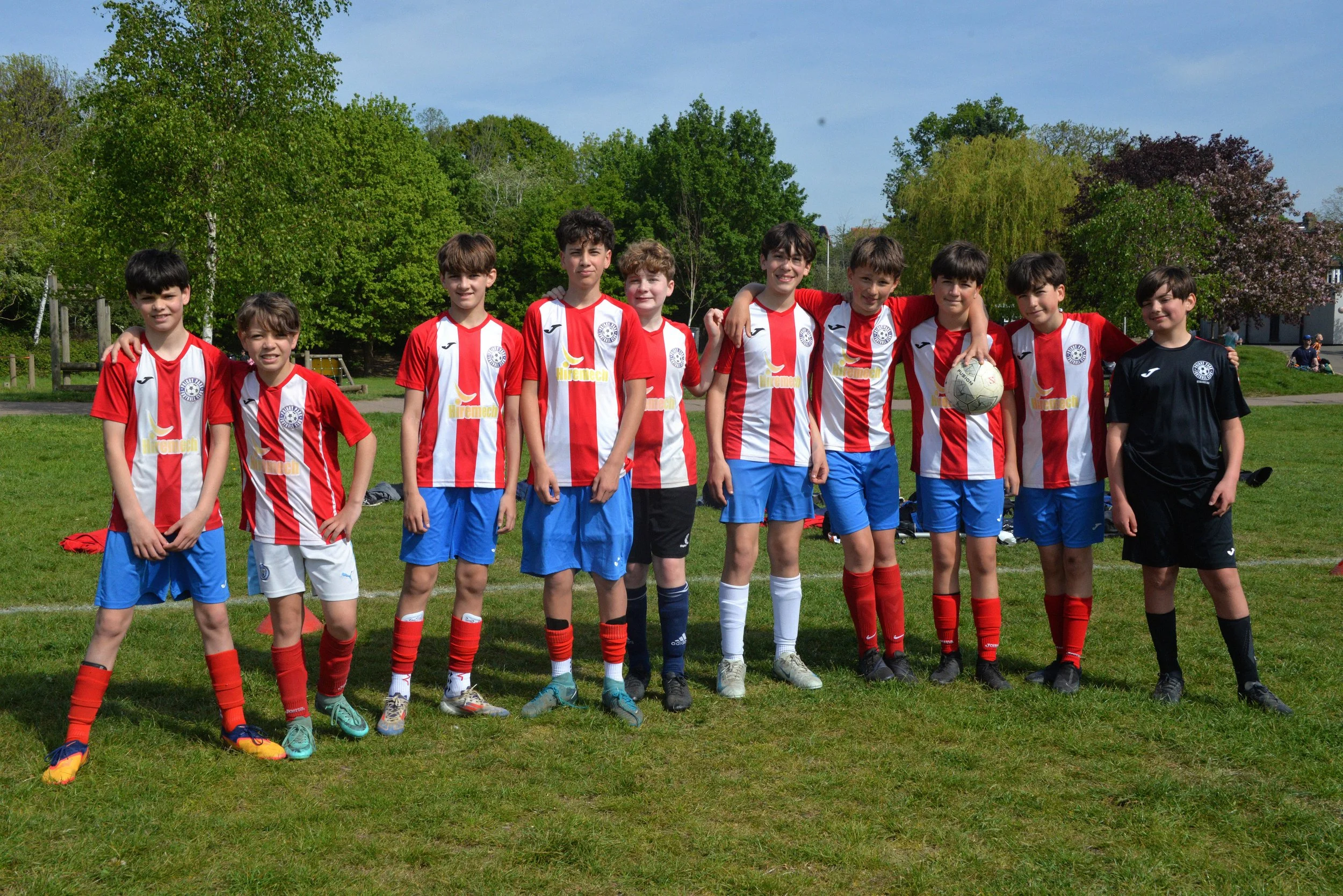 A group of young boys in soccer uniforms, standing on a grassy field with trees in the background, posing for a team photo.