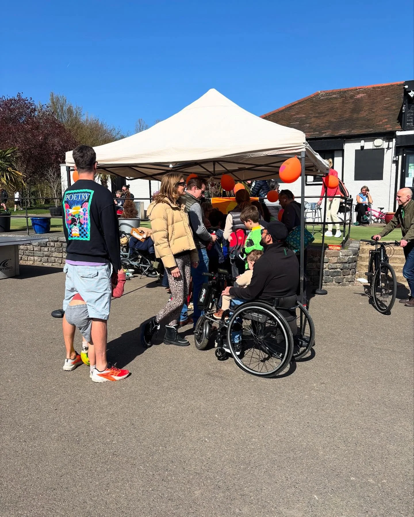 People gathered outdoors under a white canopy with orange decorations on a sunny day. Some are standing and some are in wheelchairs, engaging in conversation. Bicycles are nearby, and there is a white building with a sloped roof in the background.