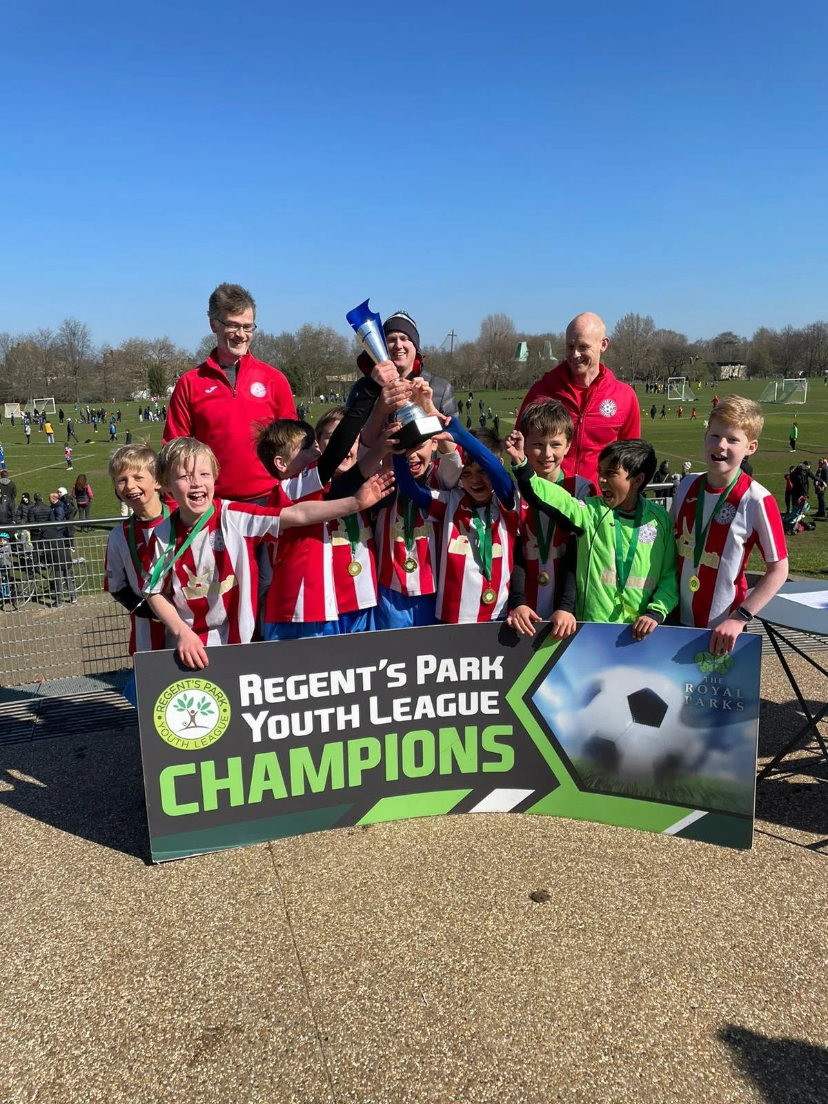 Kids in red and white striped soccer uniforms celebrating with medals and a trophy at a soccer field, holding a sign that says 'Regent's Park Youth League Champions,' with three adults behind them and a blue sky above.