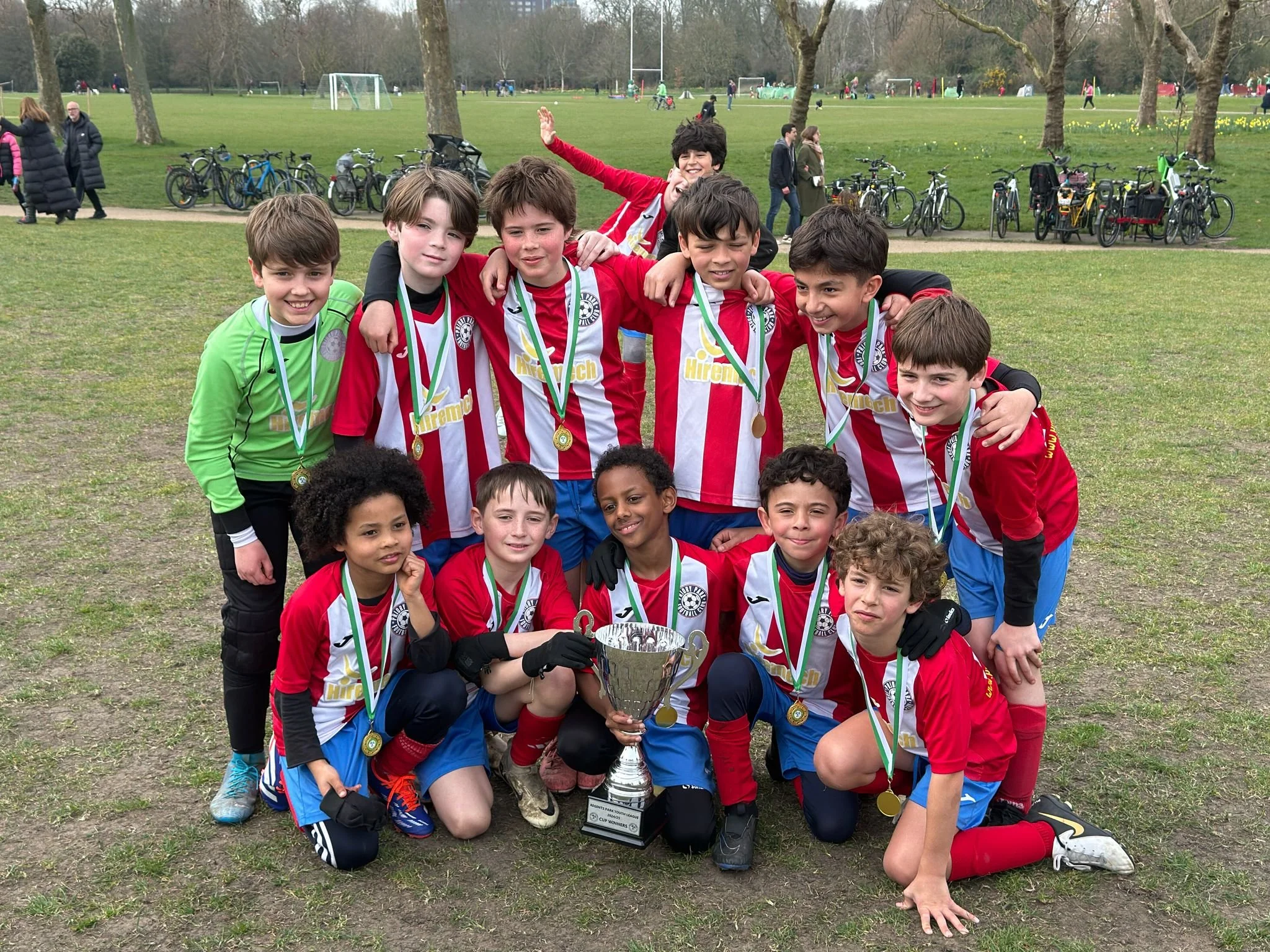 Group of young boys in red and blue soccer uniforms posing together outdoors, some with medals around their necks, holding a trophy, celebrating after winning a soccer tournament in a park with trees and bicycles in the background.