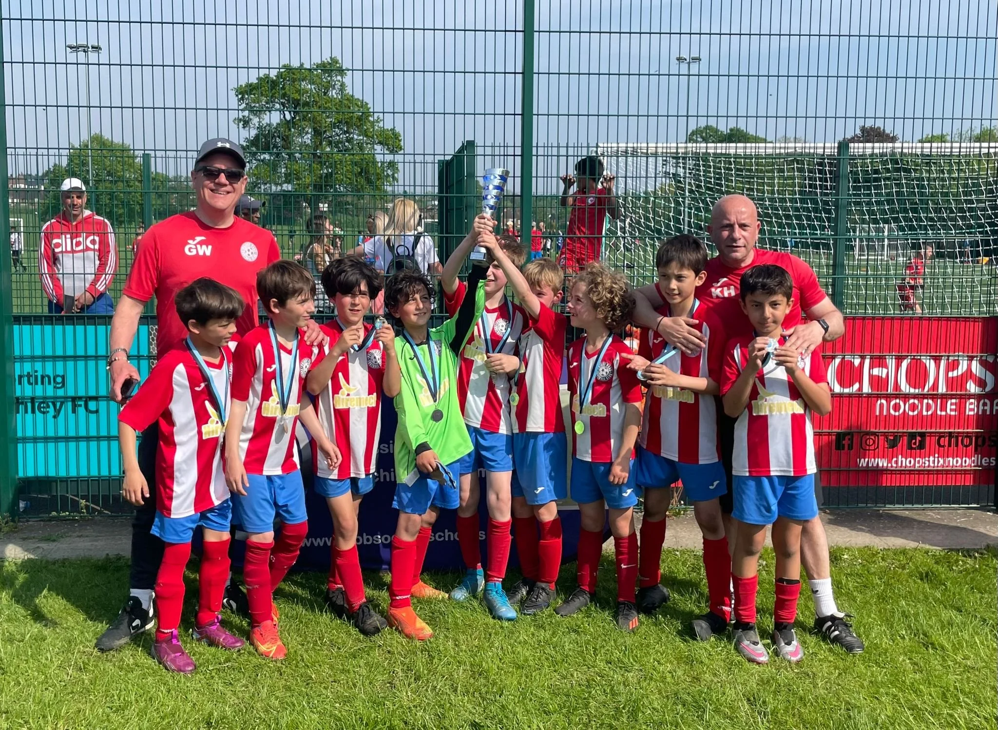 A group of young soccer players in red and white striped uniforms celebrating with medals and a trophy after a game, along with their coaches, in front of a green metal fence at an outdoor soccer field.