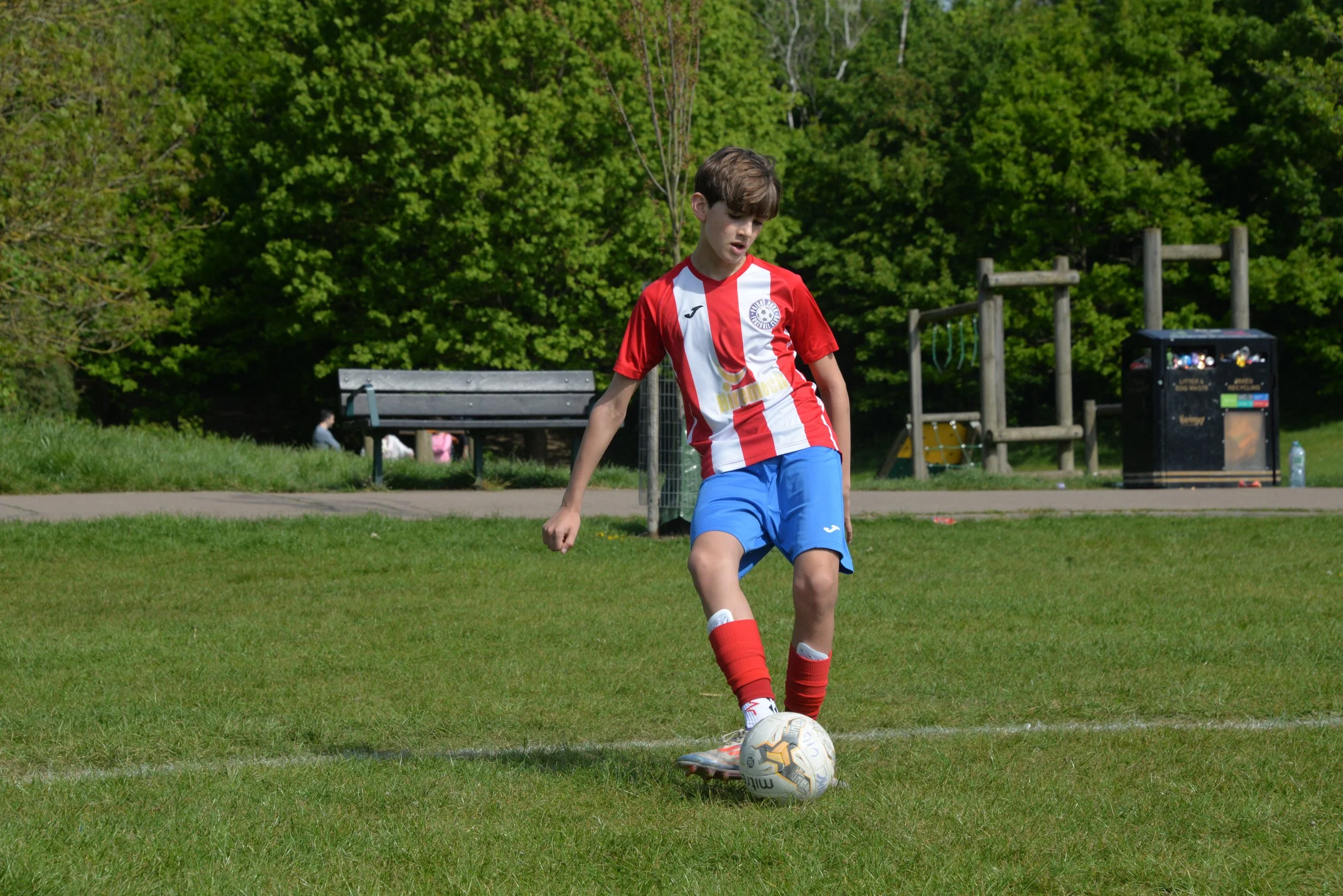 A young boy in a red and white striped soccer jersey and blue shorts is playing soccer outside on a grassy field. He is standing with one foot on a soccer ball, preparing to kick. Behind him, there is a park with benches, a trash bin, playground equi
