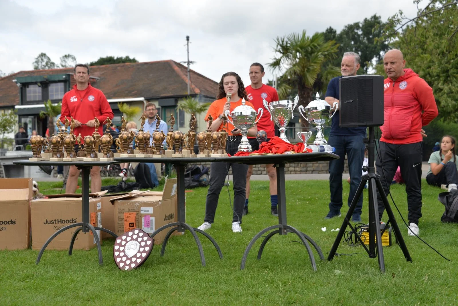 A woman in an orange shirt speaking into a microphone at an outdoor awards ceremony with trophies on a table, surrounded by men in red and blue jackets and onlookers.