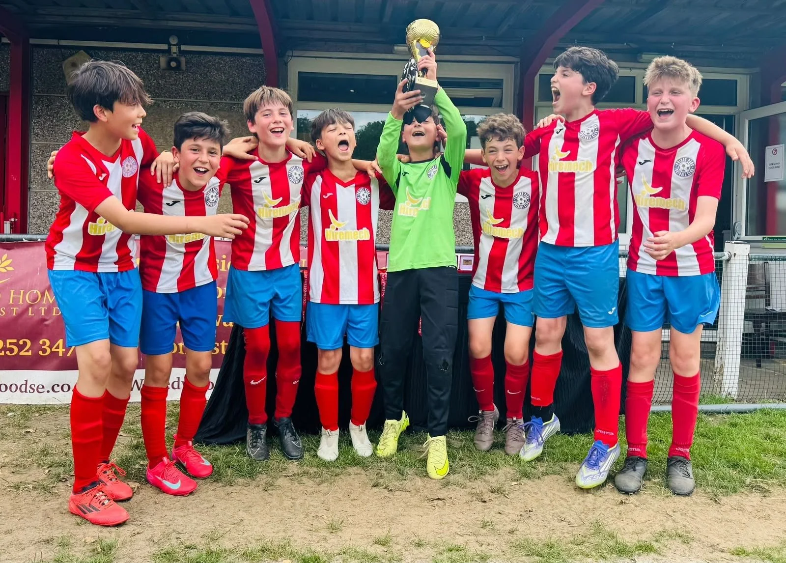 A group of young boys in red and white soccer uniforms celebrating after a match. They are standing together, smiling, with one boy in a green goalkeeper jersey holding a trophy above his head. They are on a grassy area outside a building.