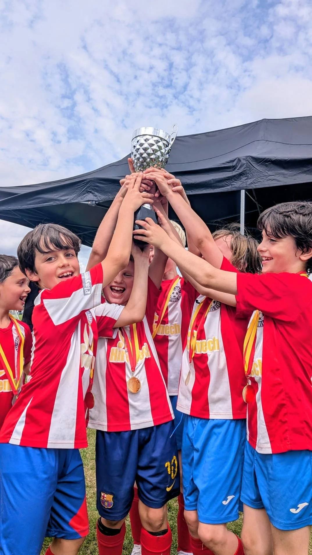 A group of young boys in soccer uniforms celebrating and holding a trophy together during daytime.