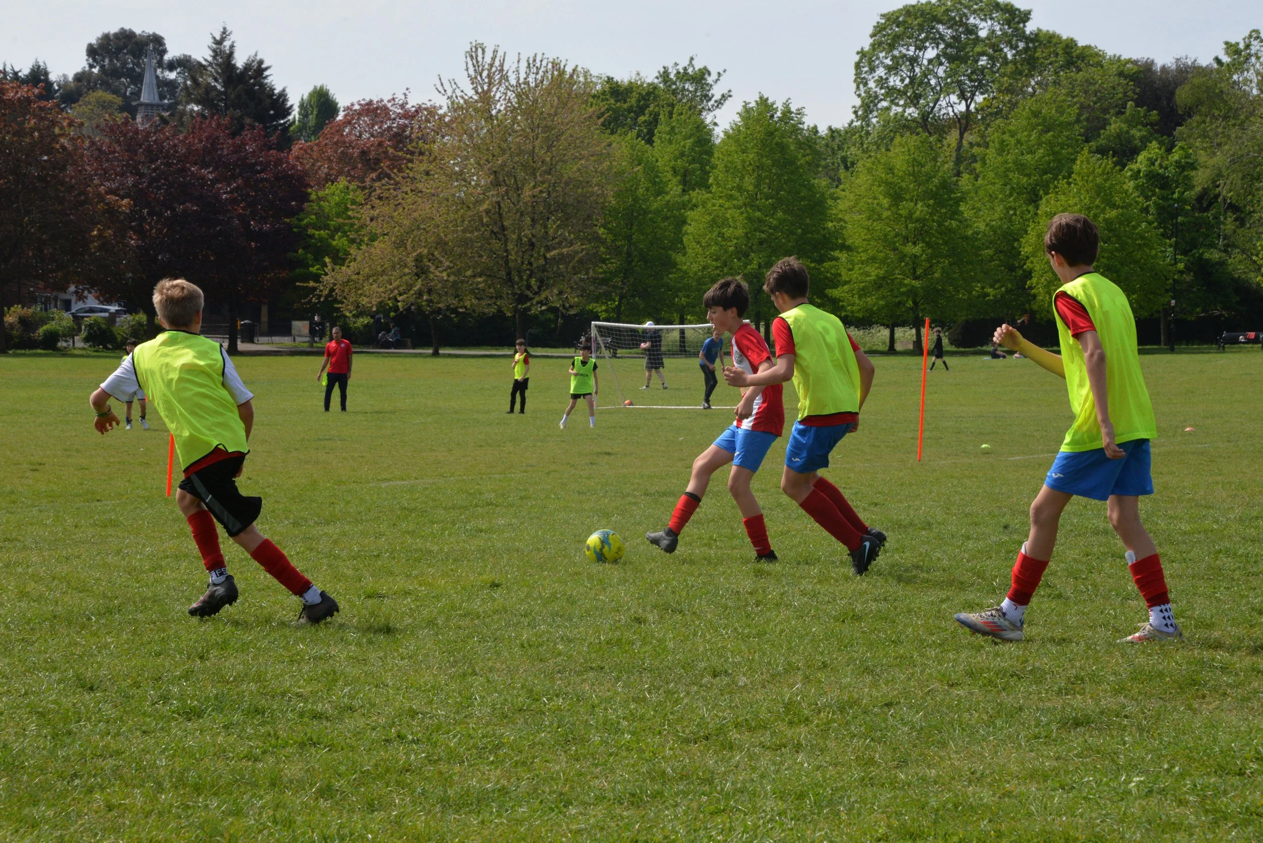 Children playing soccer on a green field in a park with trees and a few onlookers in the background.