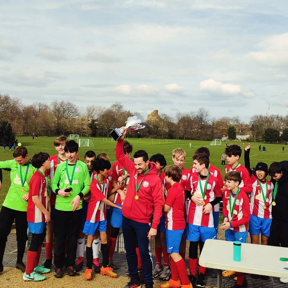 A youth soccer team in red and white jerseys celebrating with medals on a soccer field, with a man holding a trophy and smiling.