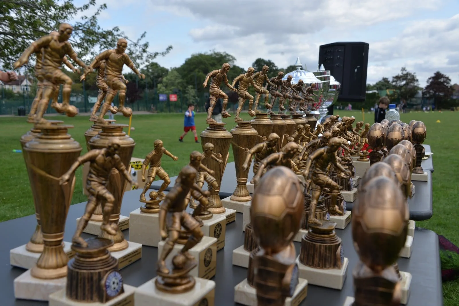 A table displaying multiple chess trophies shaped like soccer players and soccer balls, with children playing in the background on a grassy field.