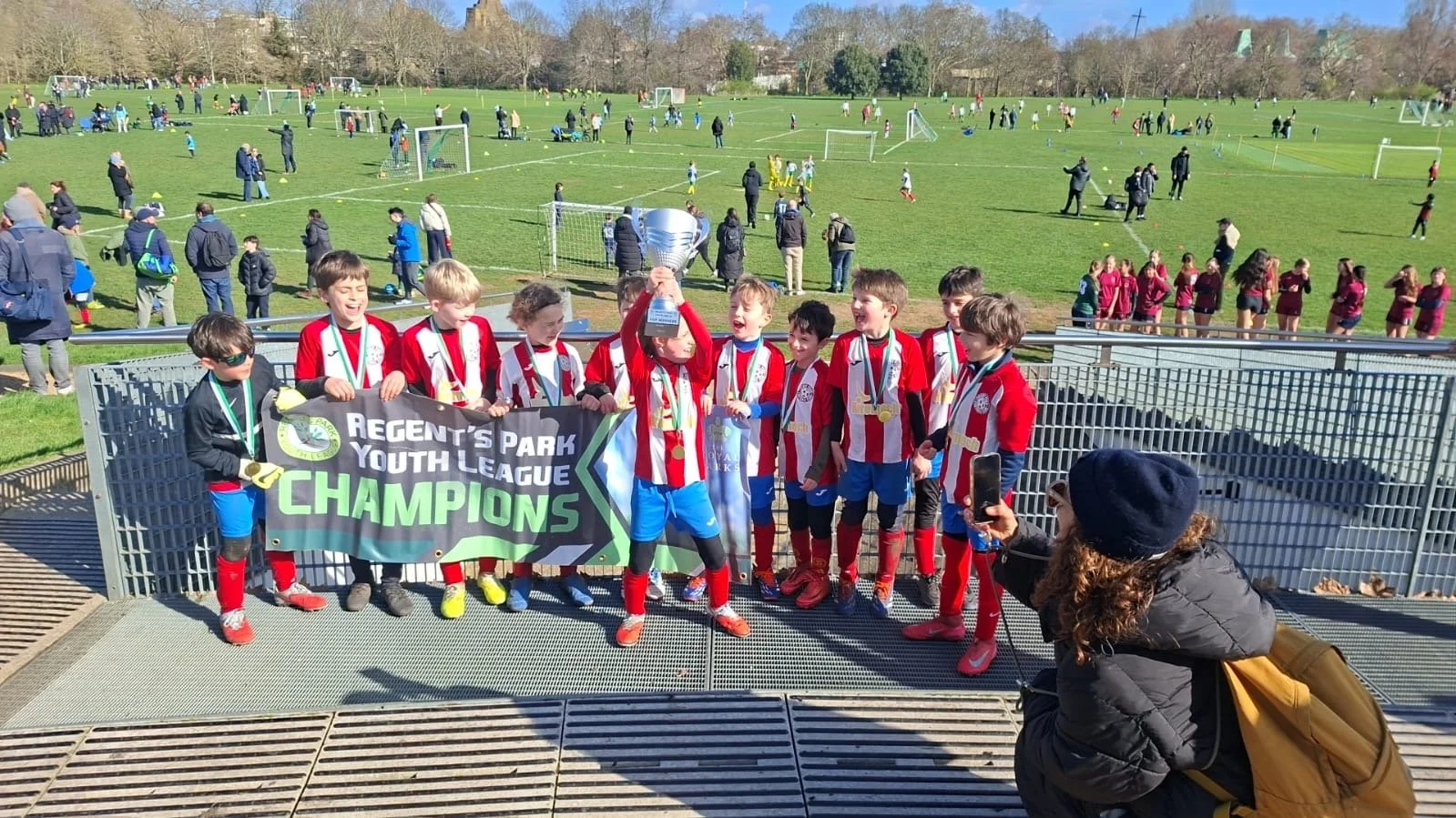 Youth soccer team celebrating winning the tournament, holding a trophy, with medals around their necks, and a banner reading 'Regent's Park Youth League Champions.' Background shows other players, fans, and soccer fields.