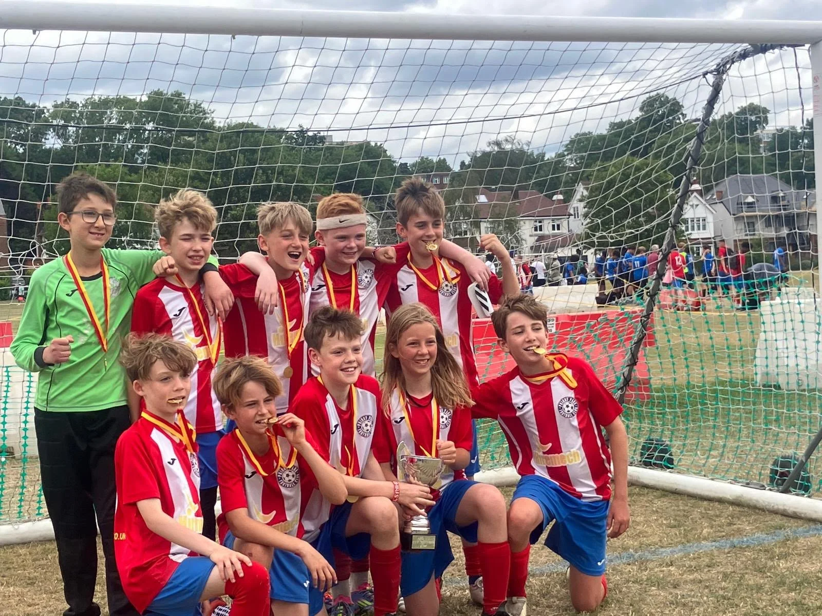 A group of young soccer players celebrating on the field after winning, wearing red and white uniforms, with medals around their necks and holding a trophy, in front of a soccer goal.