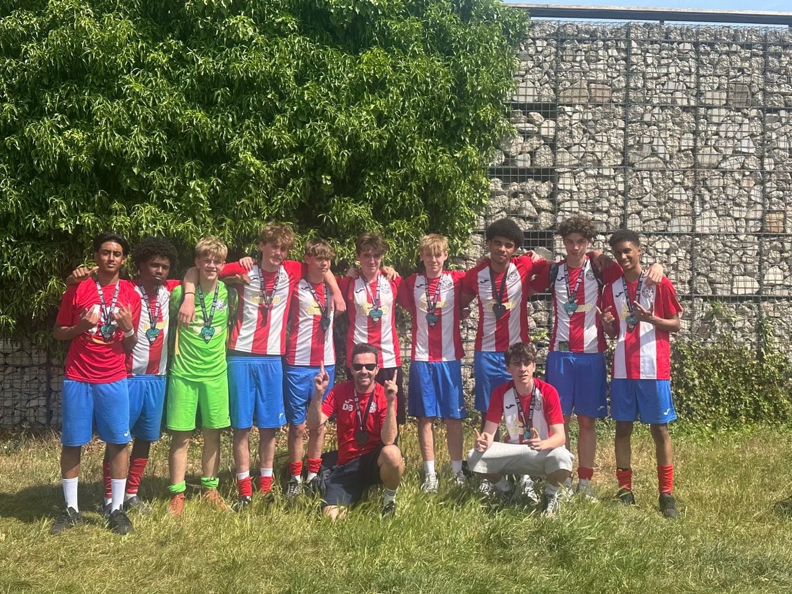 A youth soccer team and coach posing outdoors with medals after a game, wearing red and white striped jerseys and blue shorts, with a lush green bush and a rock wall in the background.