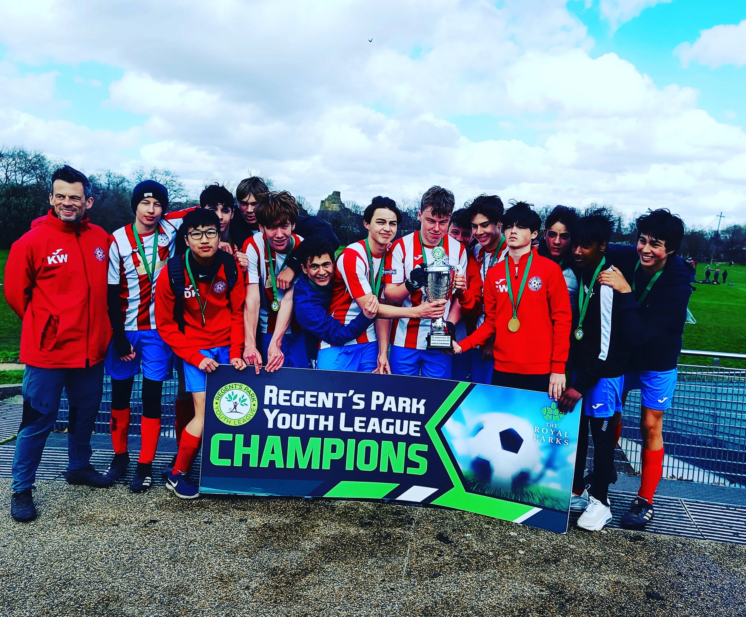 A group of young soccer players and their coach celebrating a victory at Regent's Park Youth League. They are holding a trophy and standing behind a sign that reads 'Champions,' with trees and cloudy sky in the background.
