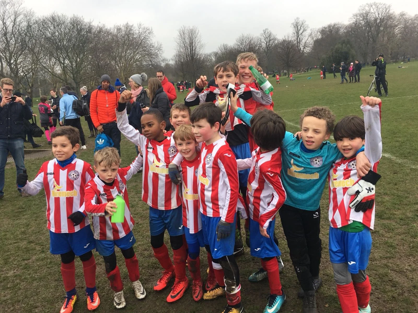 A group of young boys in red and white striped soccer jerseys celebrating on a green field. Some are posing with raised fists, and two boys at the back are holding bottles. Other kids and spectators are visible in the background on a cloudy day.