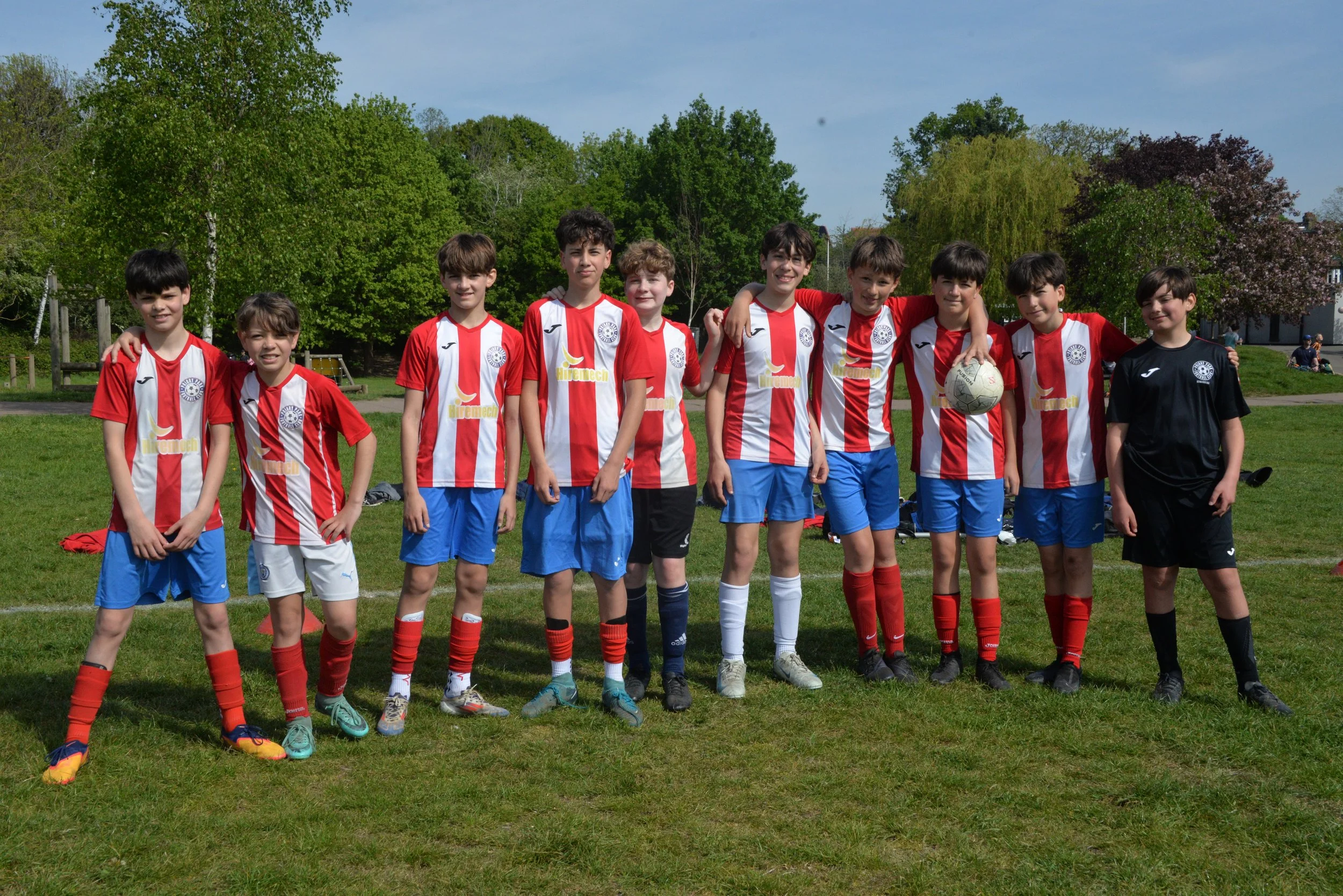 A group of young boys in soccer uniforms standing on a grassy field, posing for a photo with trees and a cloudy sky in the background.