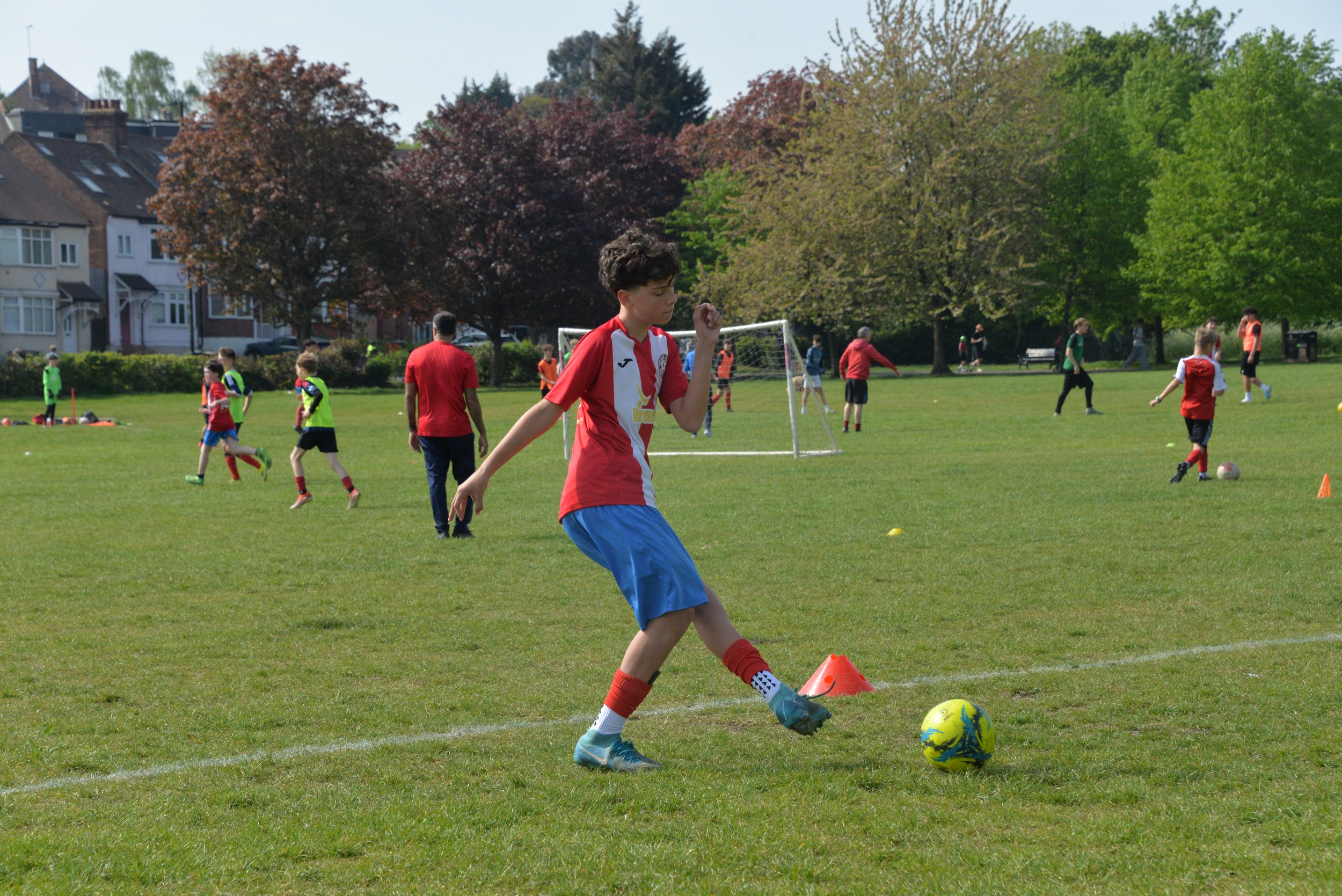 A young boy in red and blue sports attire, kicking a yellow and blue soccer ball on a grassy field during a soccer practice or game, with other children and adults in the background.