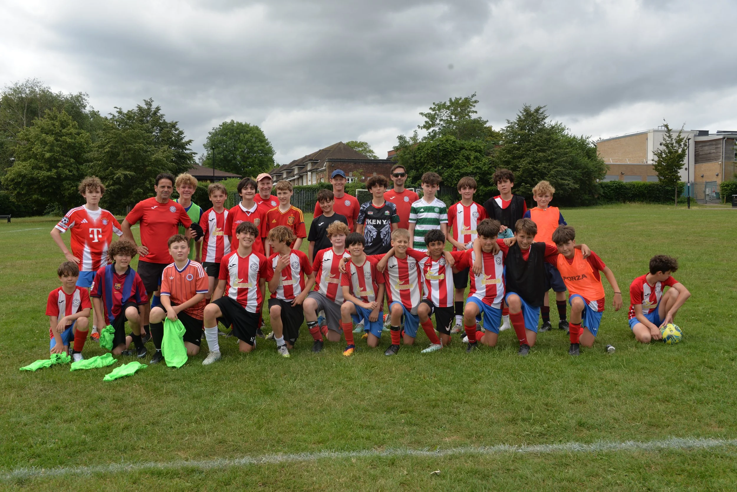 Group photo of young soccer players and coaches on a grassy field, some wearing red and white uniforms, with a row of trees and buildings in the background, under cloudy skies.
