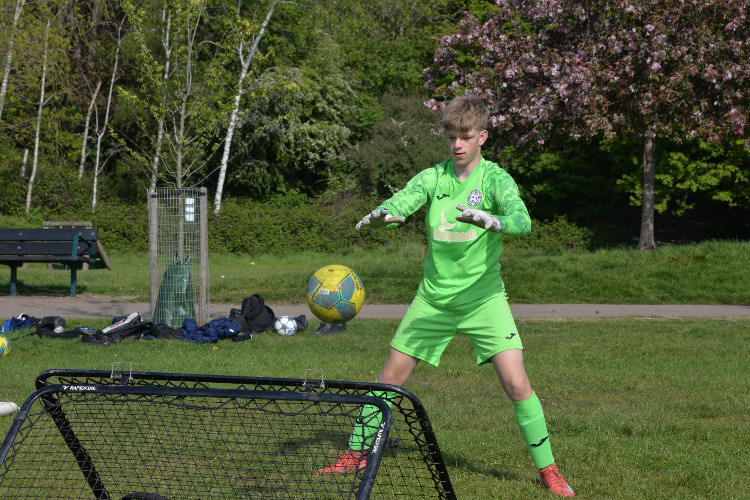 A young male soccer player in a bright green uniform practicing soccer outdoors on a grassy field with trees and pink blossoms in the background.