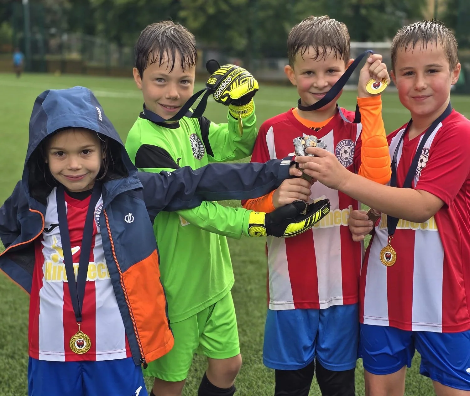 Young soccer players celebrating with medals and a trophy on a soccer field after a match, with a girl wearing a rain jacket on the left.