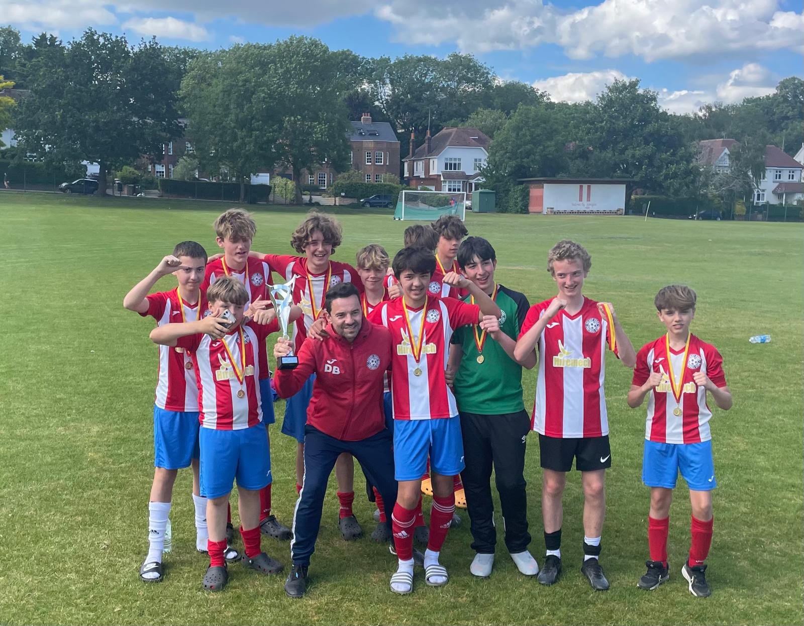 Youth soccer team in red and white jerseys celebrating wins on a grassy field, holding medals and a trophy, with trees and houses in the background under a partly cloudy sky.