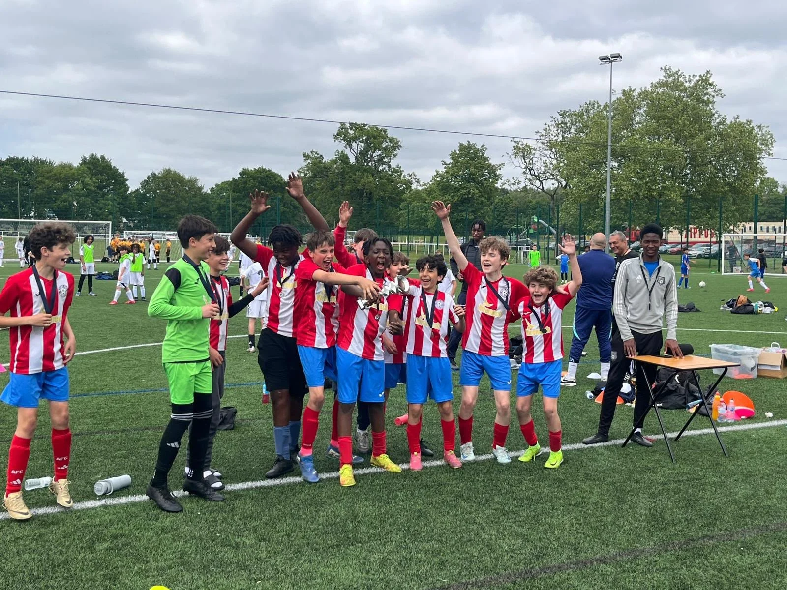 A group of young soccer players celebrating on a field, with some raising their hands and holding a trophy, while others smile and cheer. They are dressed in red and white striped jerseys and blue shorts. In the background, other players and coaches 