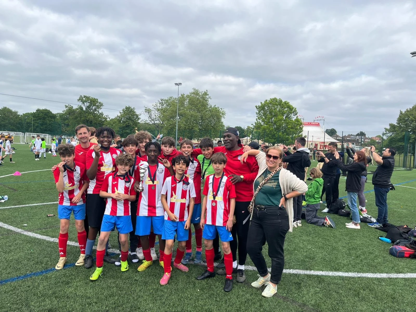 A youth soccer team with their coaches and supporters, celebrating with medals and a trophy on a soccer field.
