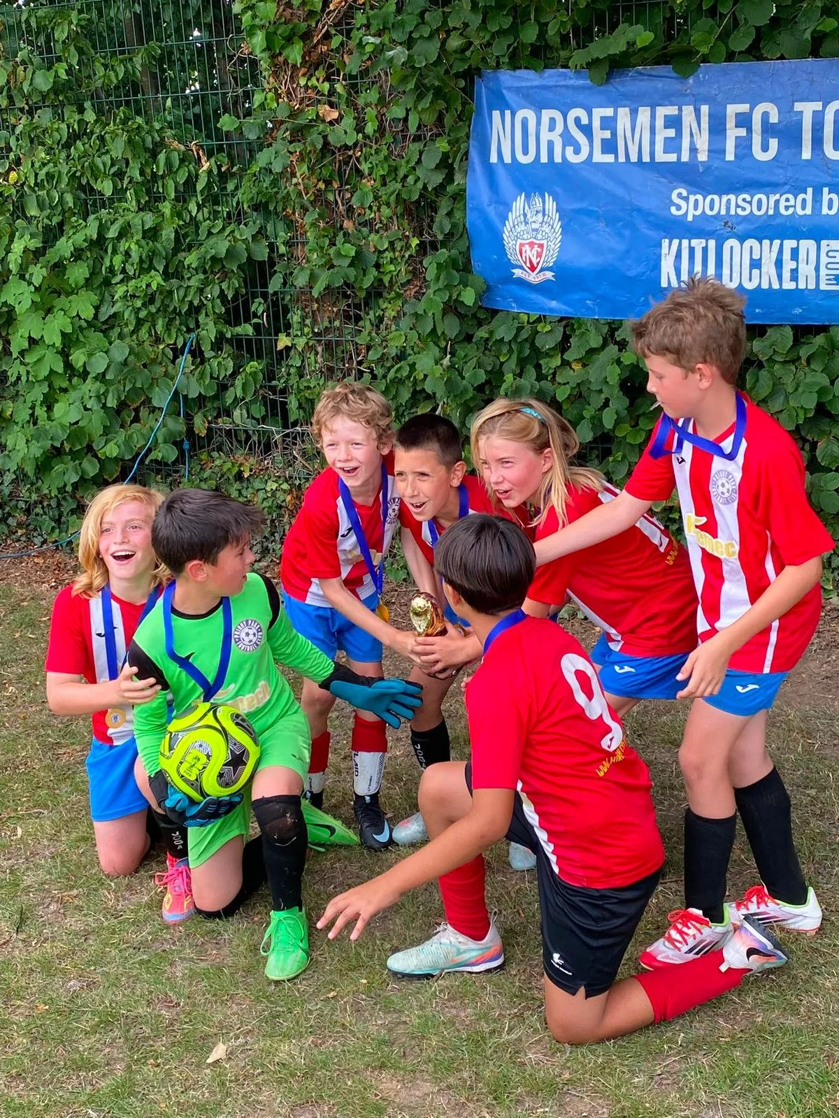 Kids in red and blue soccer uniforms celebrating with a trophy, with some kneeling and others standing, in front of green bushes and a blue banner that reads 'Norsemen FC'.