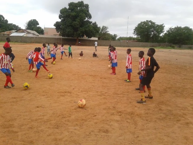 Children playing soccer on a dirt field with some trees and buildings in the background.