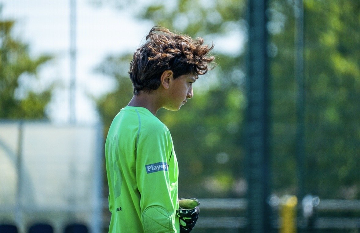 Young soccer goalkeeper in bright green jersey with glove, standing on a soccer field, looking down thoughtfully during daytime.