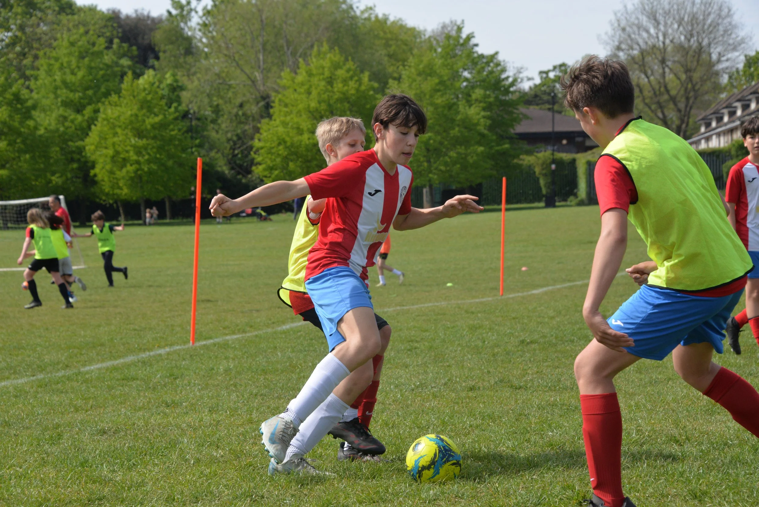 Children playing soccer on a grassy field with trees and a building in the background.