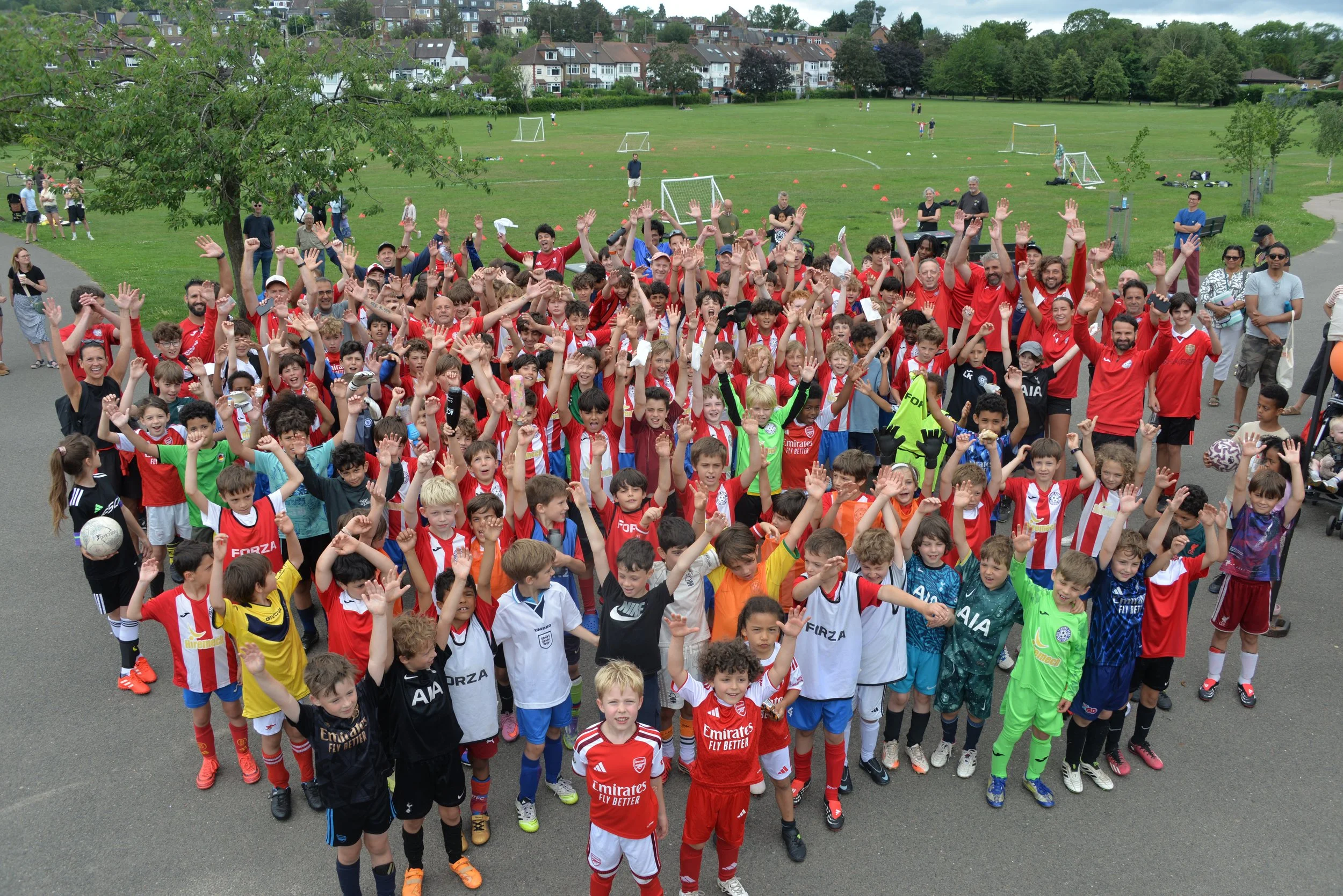 A large group of children and adults gathered outdoors in a park, many wearing colorful sports jerseys, waving, and smiling for a group photo with soccer fields and trees in the background.