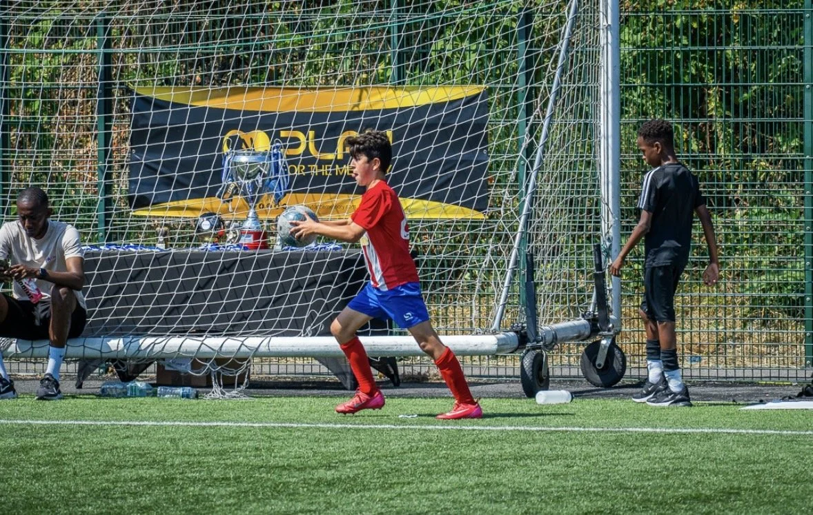 Children playing small-sided soccer game with goal, one child in red uniform holding a soccer ball, another child in black uniform near the goal, and a boy sitting on the ground.