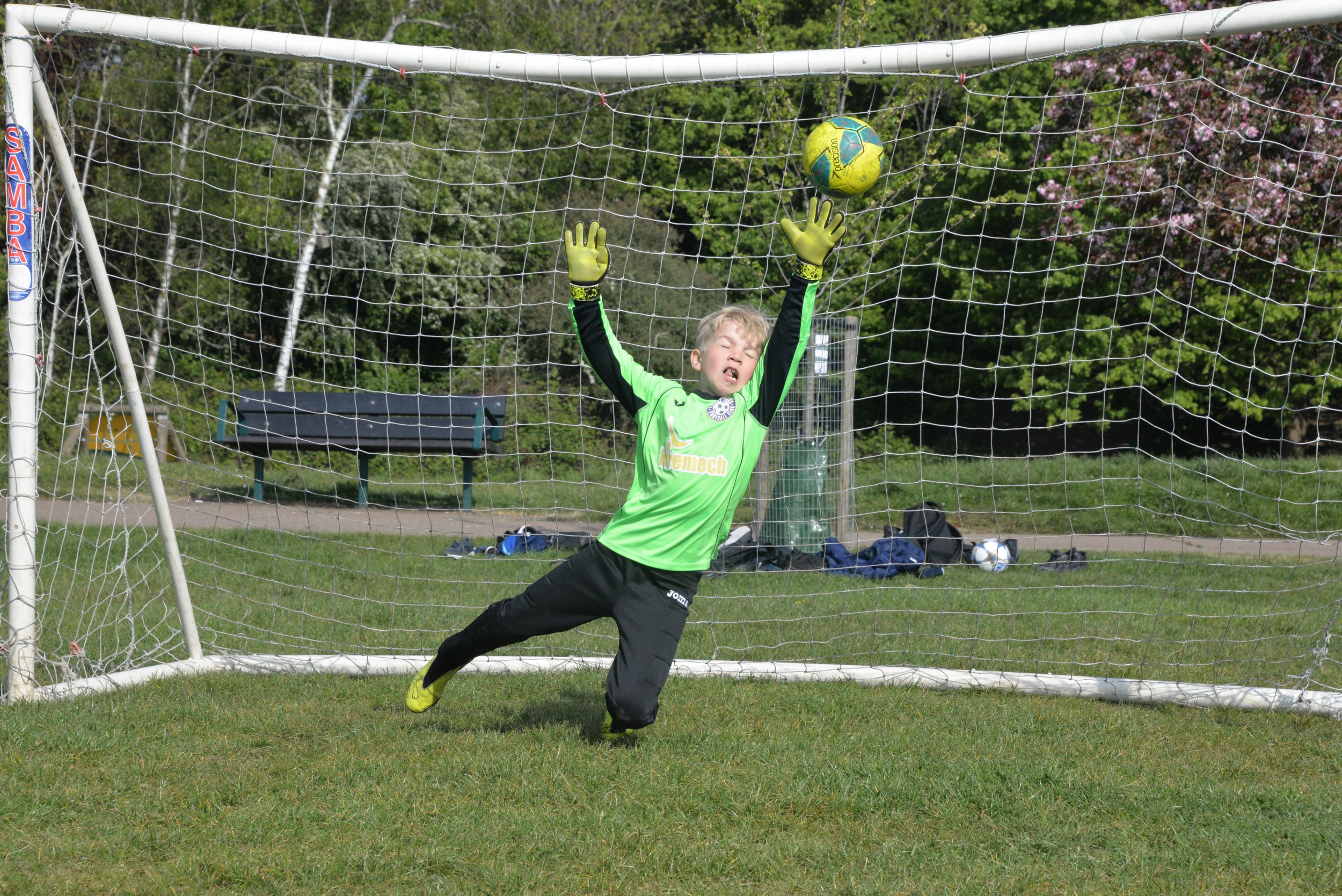 A young boy playing soccer as a goalkeeper, jumping and reaching with both hands to block a bright yellow and green soccer ball near the goal on a grassy field, with trees and benches in the background.