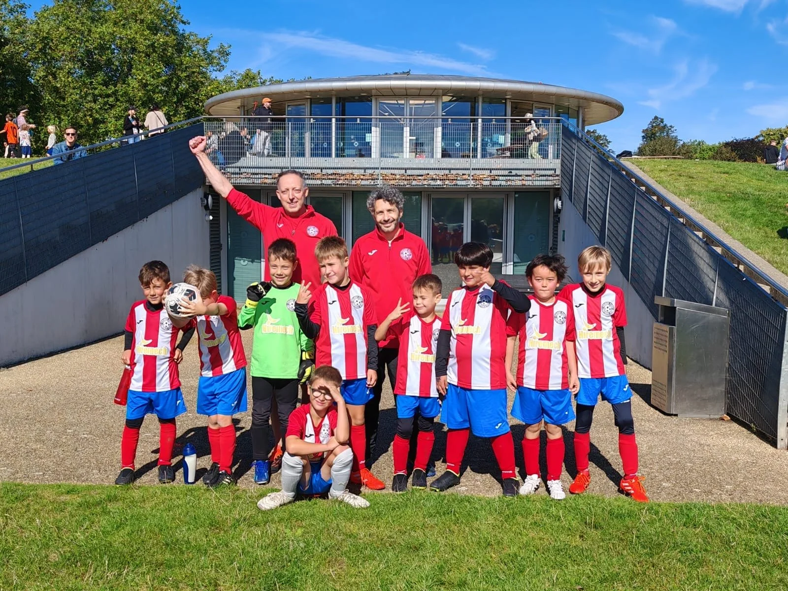 Group of young soccer players and coaches pose for a photo outdoors on a sunny day, with children wearing red and white striped jerseys and blue shorts, standing on a concrete pathway against a backdrop of a building and grassy hill.