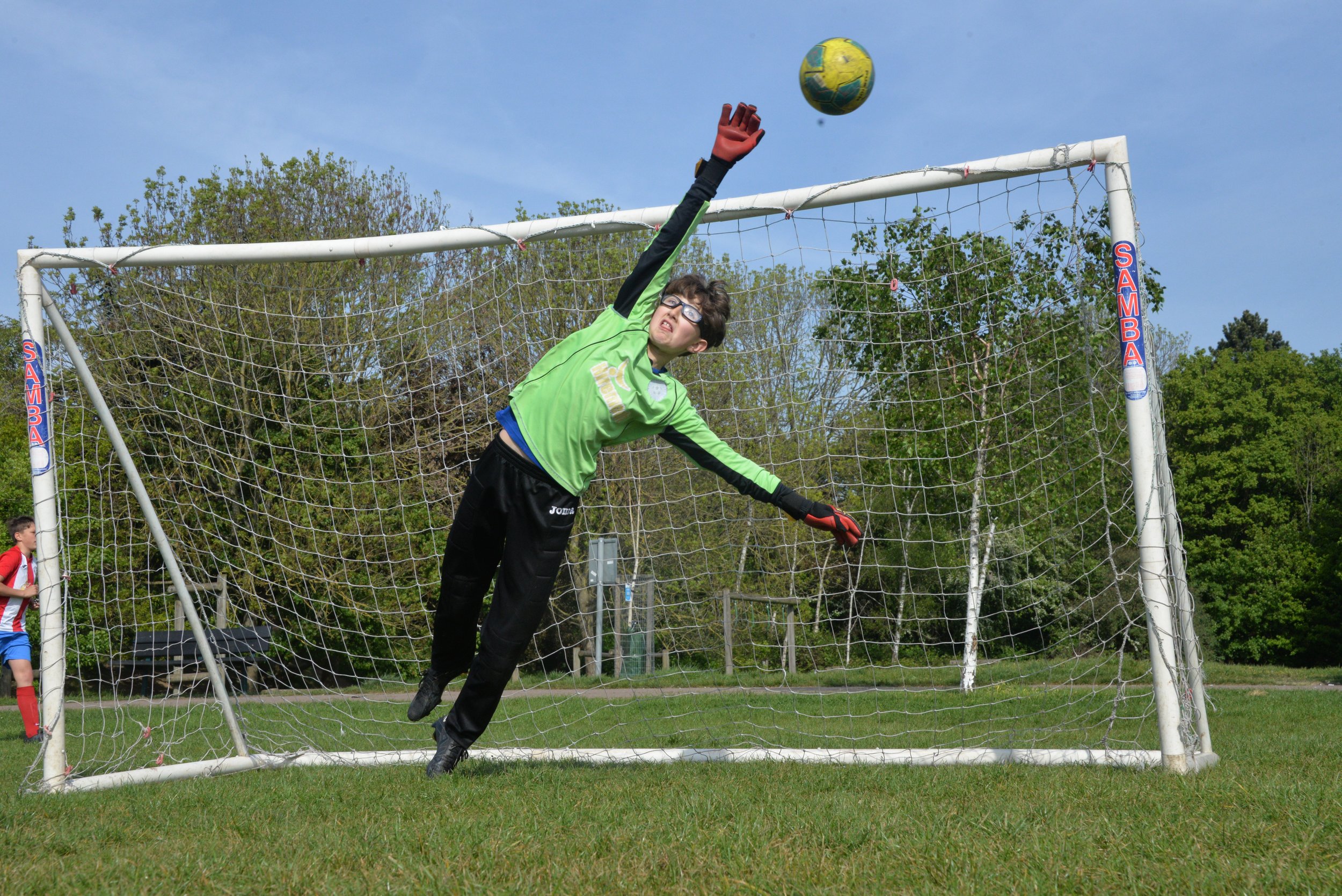 A young soccer goalkeeper wearing glasses, a green jersey, black pants, and gloves, reaching up to catch a yellow and green soccer ball during a game on a grassy field with trees in the background.