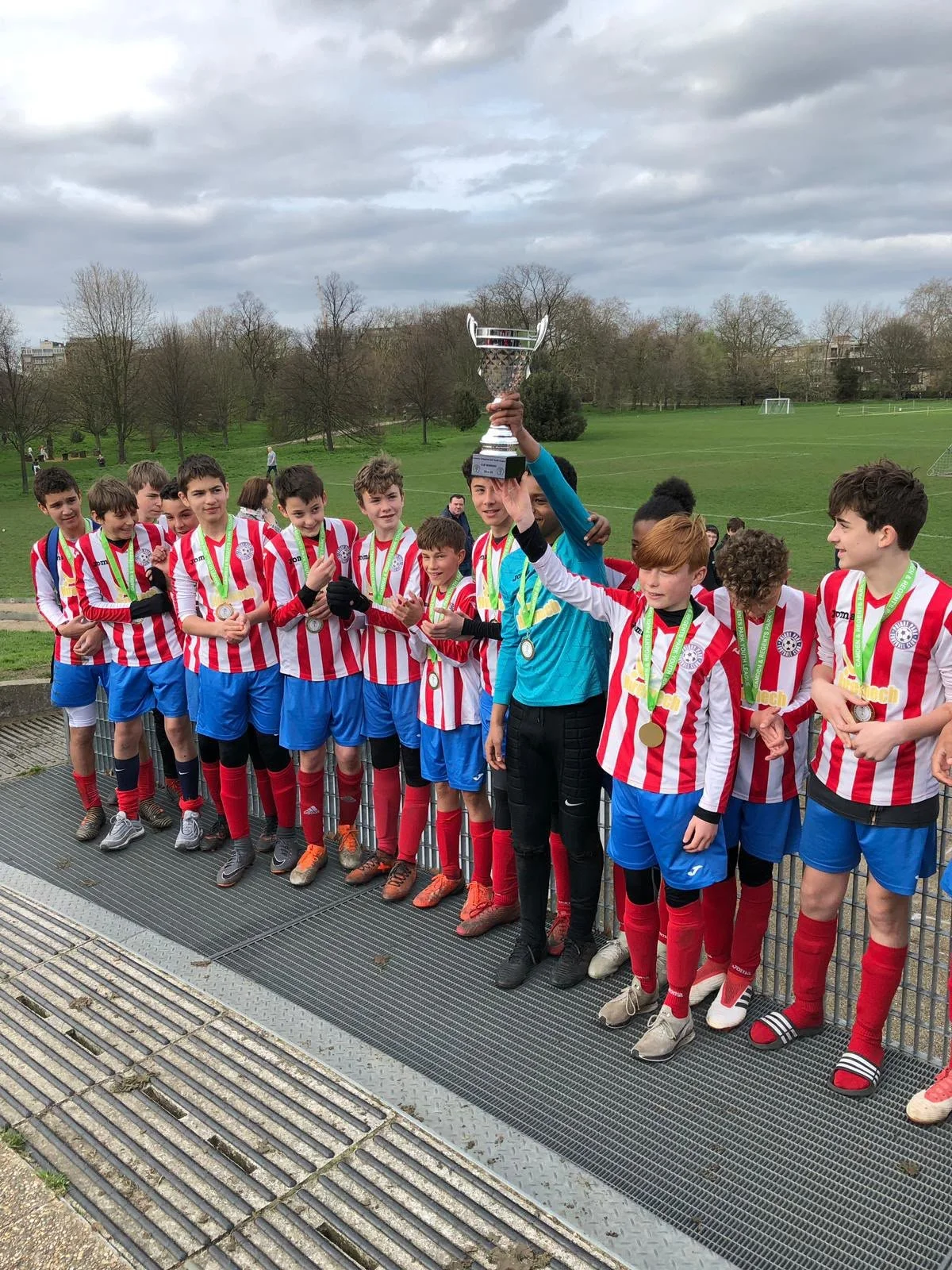 A group of young soccer players in red and white striped jerseys and blue shorts celebrating their victory on a soccer field. One of the players, wearing a blue goalkeeper jersey, is holding a large trophy above his head. The players are wearing meda