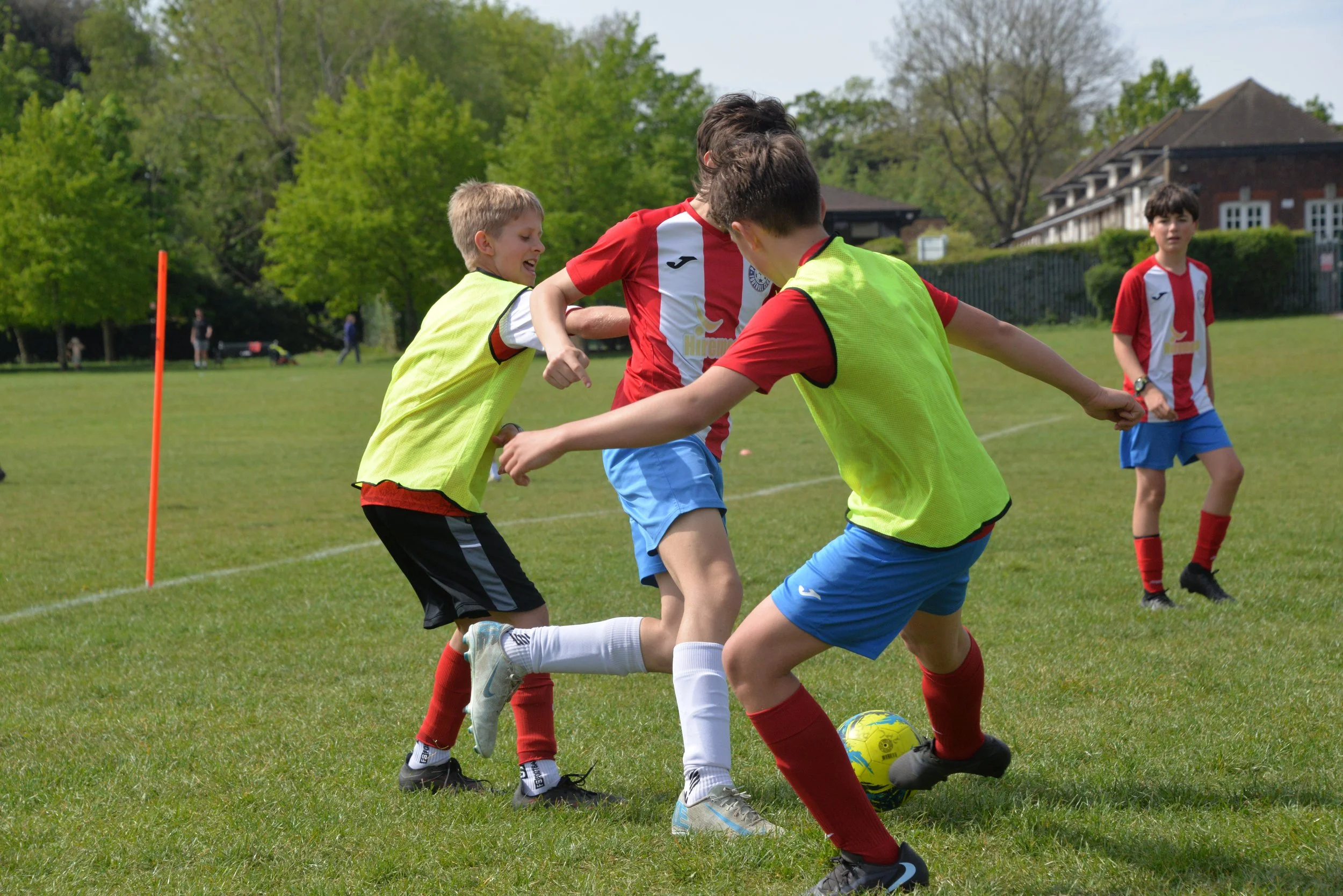 Children playing soccer on a grassy field during daytime, with trees and houses in the background.