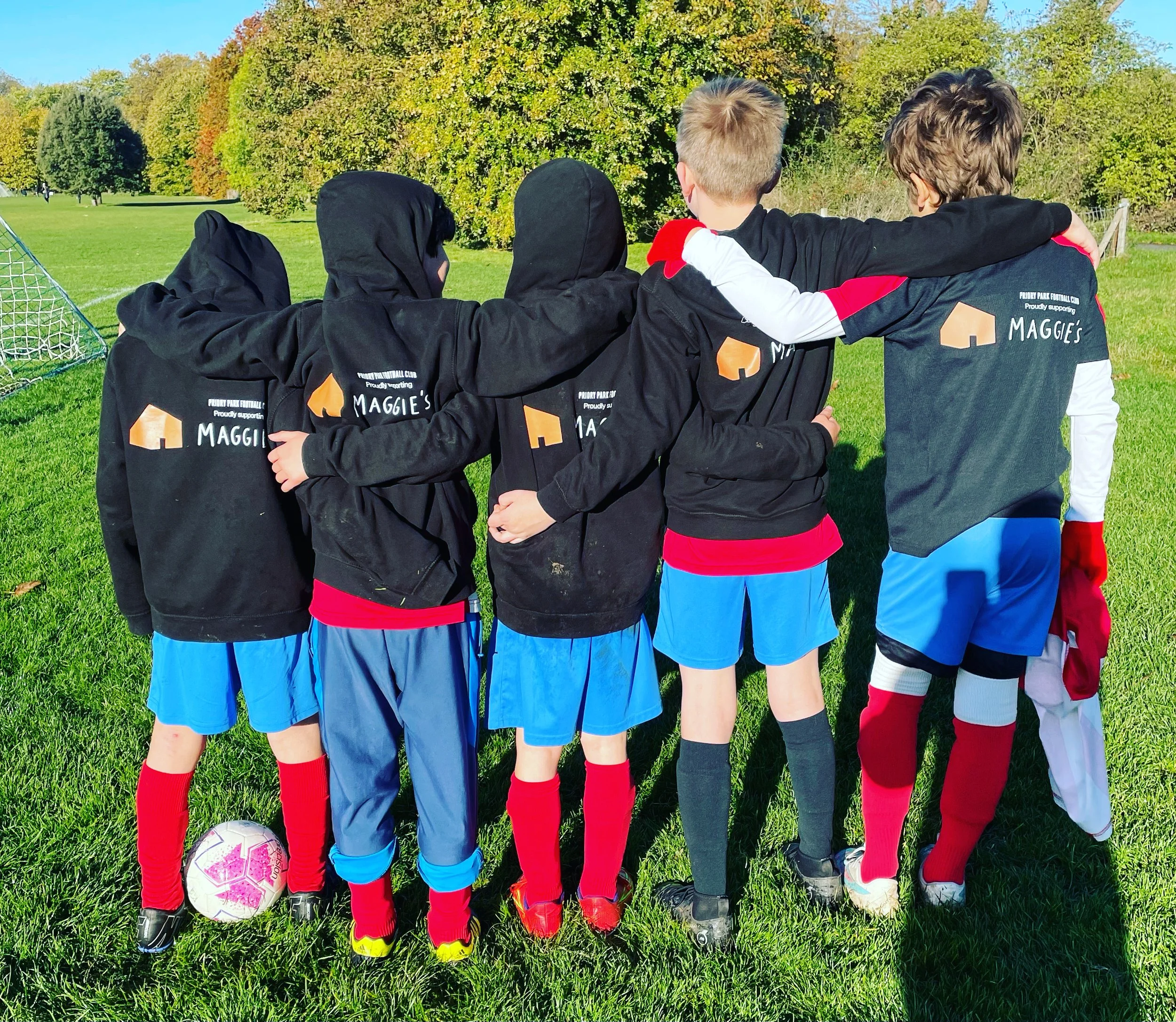 A group of children in soccer uniforms standing on a grassy field with their arms around each other, facing a soccer goal, with trees and blue sky in the background.
