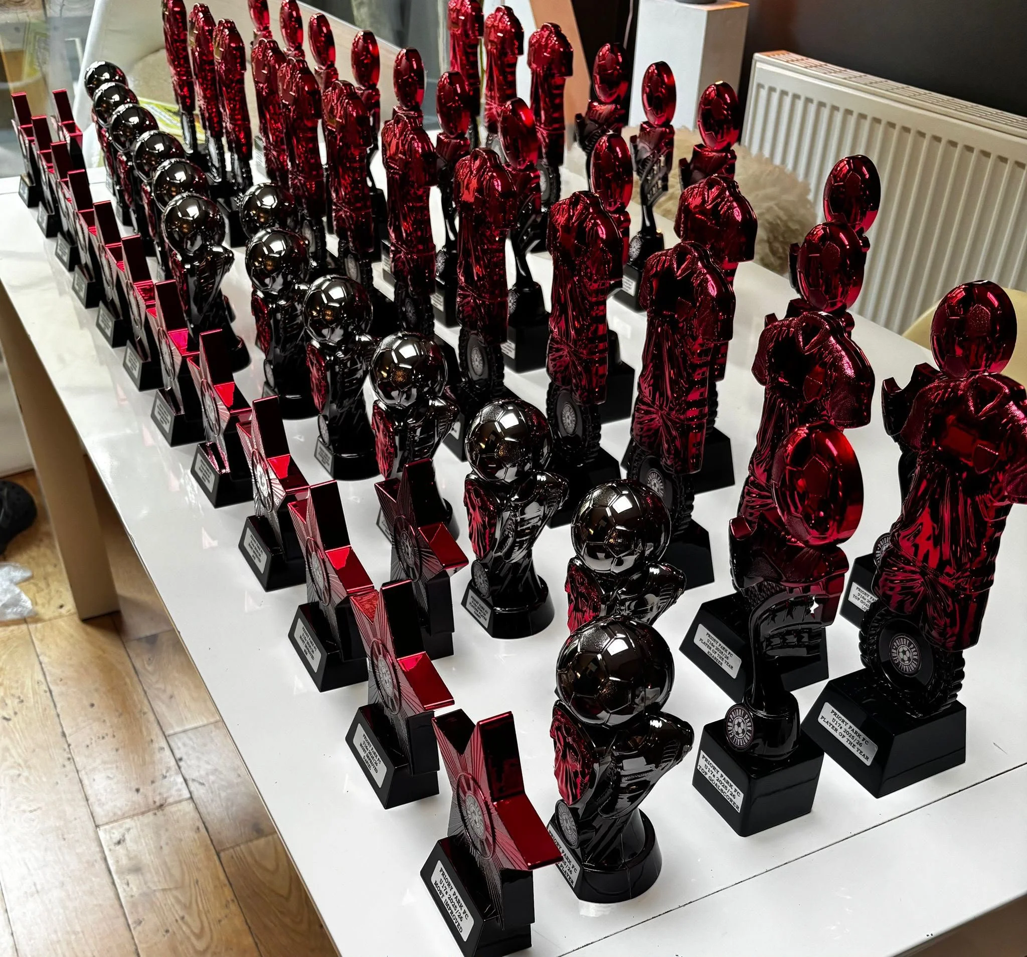 Display of multiple shiny red and black trophies shaped like human figures, arranged on black bases with labels, on a white table.