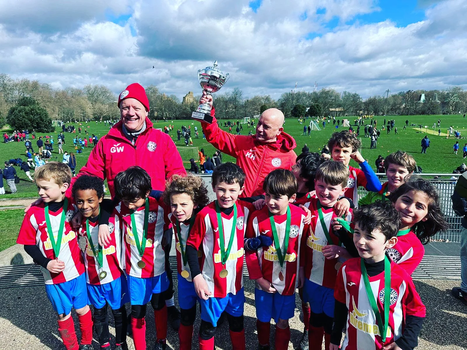Group of young children in soccer uniforms with medals, two adults in red jackets, one waving a trophy, celebrating outdoors on a cloudy day.
