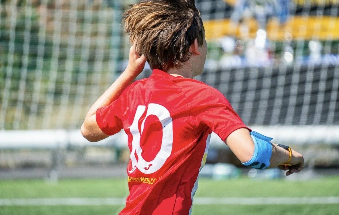 A young soccer player in a red jersey with the number 10 on the back, adjusting their hair on the field near a goal.